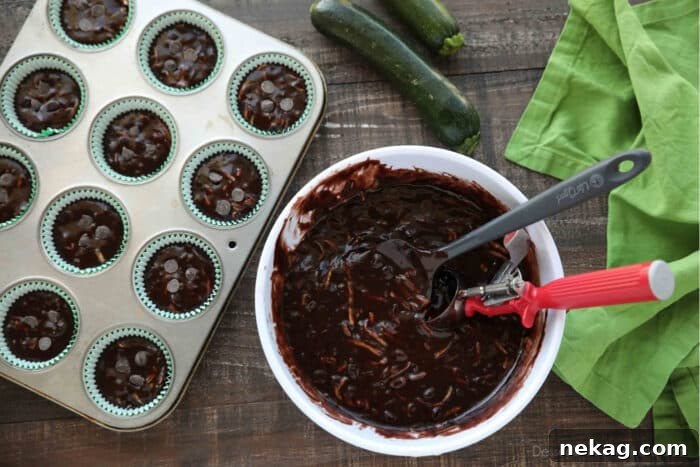 Moist Chocolate Zucchini Muffins with Melted Chocolate Chips 9 Chocolate Zucchini Muffin batter in a bowl with a spatula and scoop next to a muffin tray with paper liners and batter scooped inside.