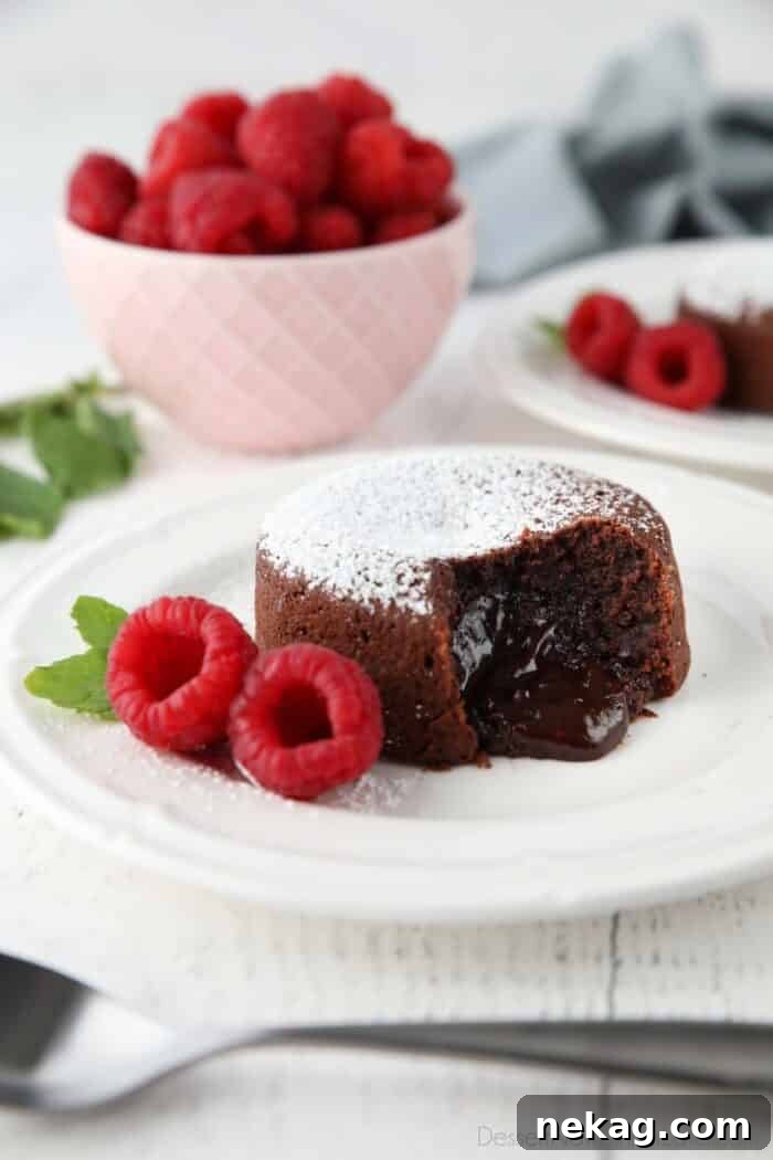 Molten Chocolate Lava Cake Recipe with Step-by-Step Video 2 Molten chocolate lava cake on plate dusted with powdered sugar and a side of raspberries and mint, with gooey center melting onto the plate.