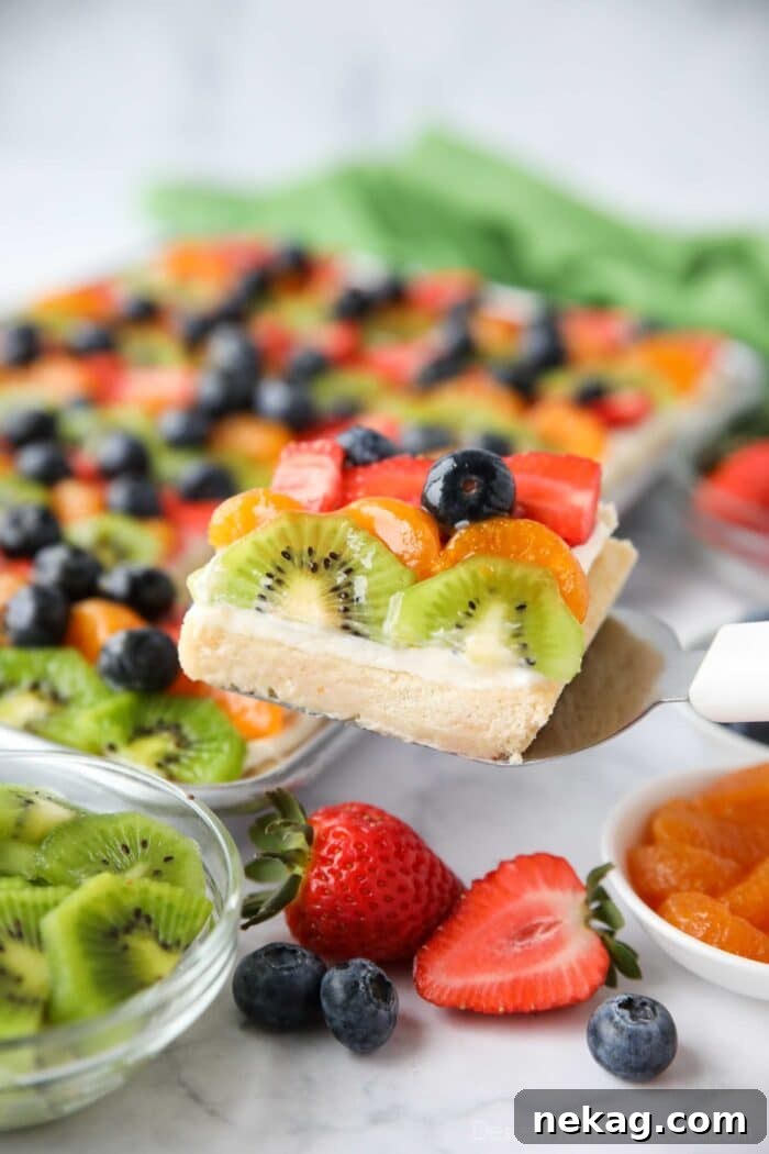 Scooping a sugar cookie fruit pizza bar with cream cheese frosting from tray.
