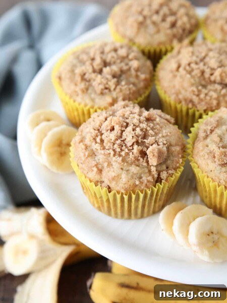 White cake plate with banana muffins in yellow wrappers with crumb streusel on top and slices of banana on the sides.