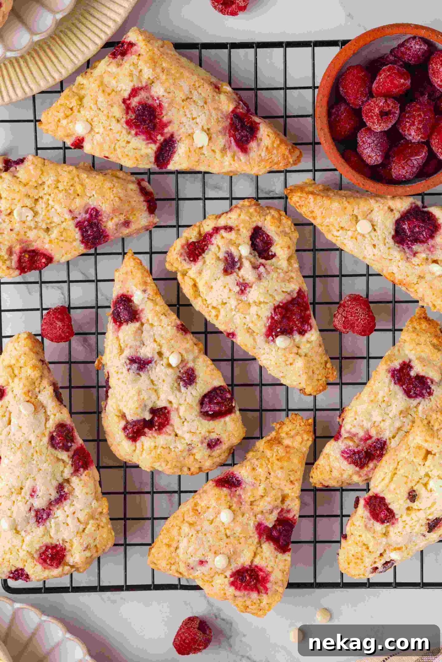 Raspberry white chocolate scones on a wire cooling rack beside a small bowl of frozen raspberries.